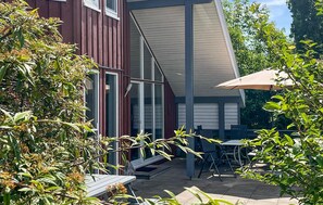 Zona de pati amb taula i cadires, espai de seients cobert i edifici amb finestres grans i façana vermella.