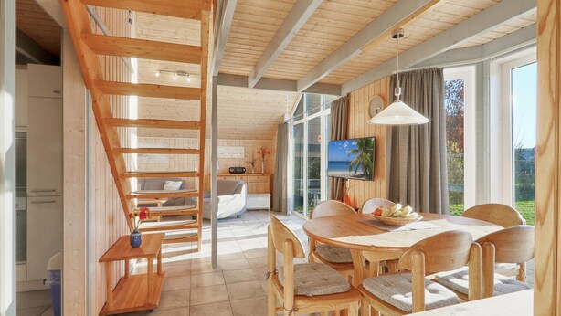 L’intérieur chaleureux d’un chalet, avec un escalier en bois, un coin repas comprenant une table et des chaises, un téléviseur et une grande fenêtre donnant sur le paysage extérieur.