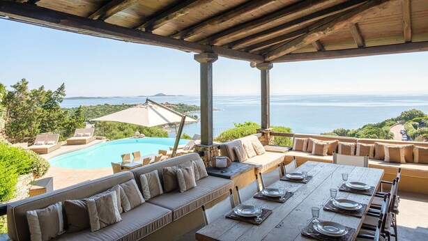 Een overdekt terras met een zwembad, een eettafel en uitzicht op zee.