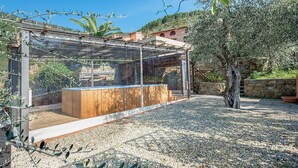 Un espace extérieur moderne aménagé avec un banc en bois et une structure en verre, entouré d’oliviers et bordé d’un chemin de gravier.