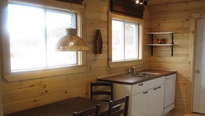 A compact kitchen with wooden walls, a dining table with chairs, a hanging light fixture, and a window with blinds.