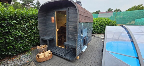 A sauna with a dark exterior, a glass door, and a chimney. There are wooden benches and baskets outside. - Quiet, centrally located vacation home with a sauna, pool, and hot tub (Wendisch Rietz)