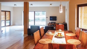 Un comedor con una mesa de madera dispuesta para cuatro personas, rodeada de sillas, con un televisor y altavoces al fondo.