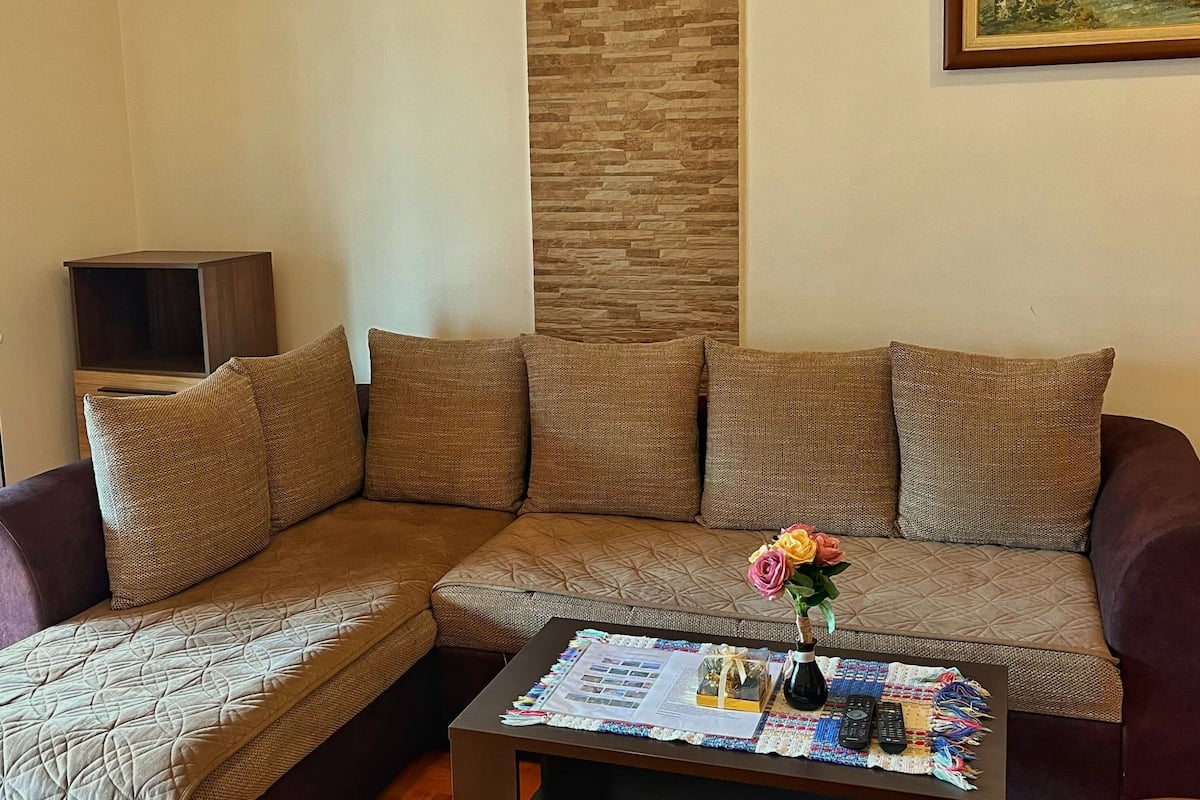 Uma sala de estar com sofá seccional, uma mesinha de centro com um vaso de flores, um quadro na parede e uma parede de detalhes em pedra.