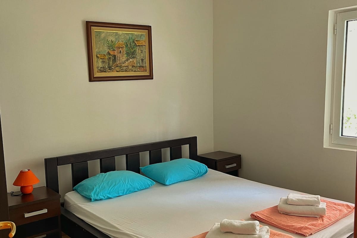 Quarto com cama, almofadas, mesa de cabeceira, abajur, pintura na parede e janela.