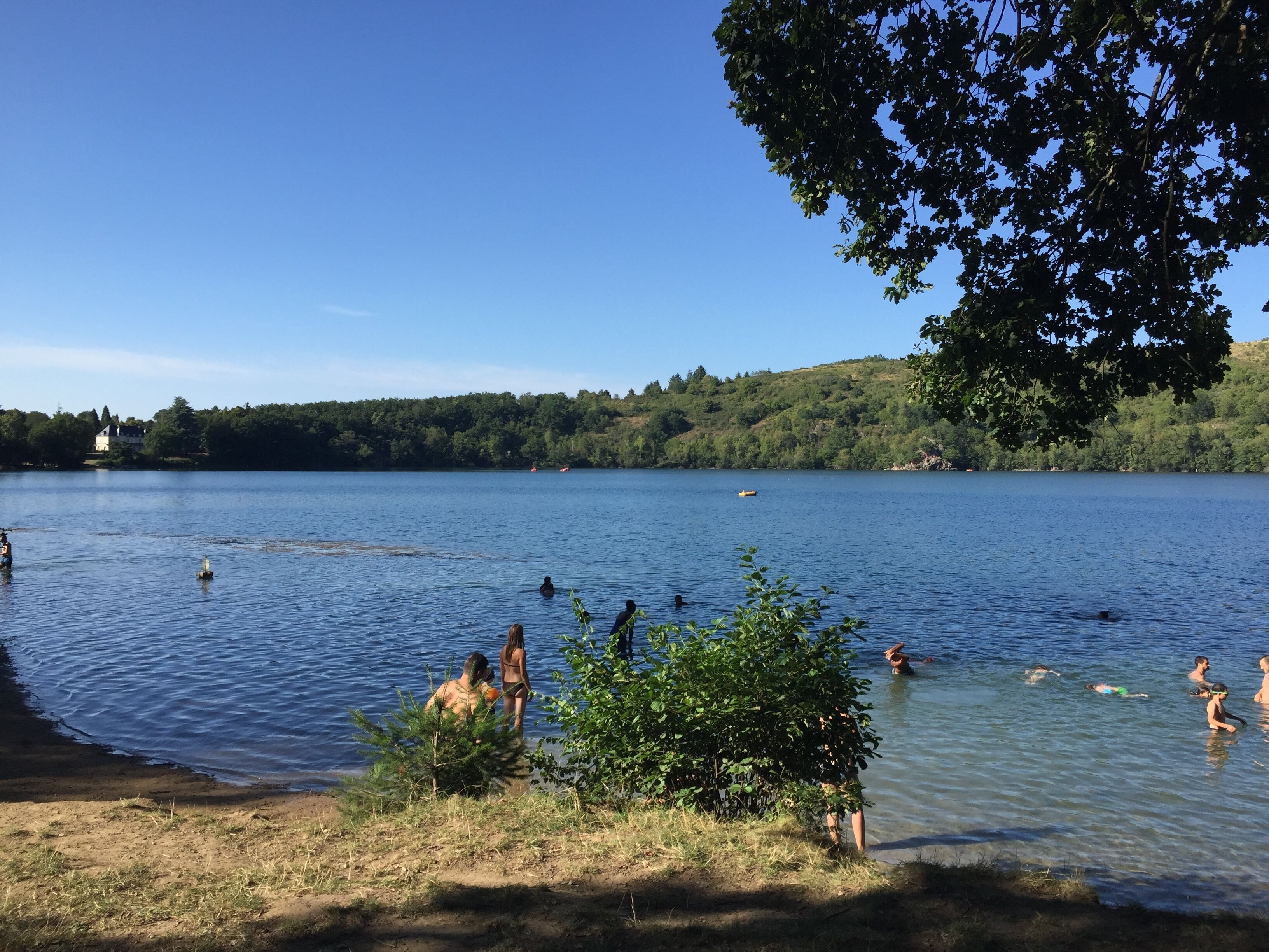 En sjö där folk badar och står på stranden, omgiven av träd och kullar.