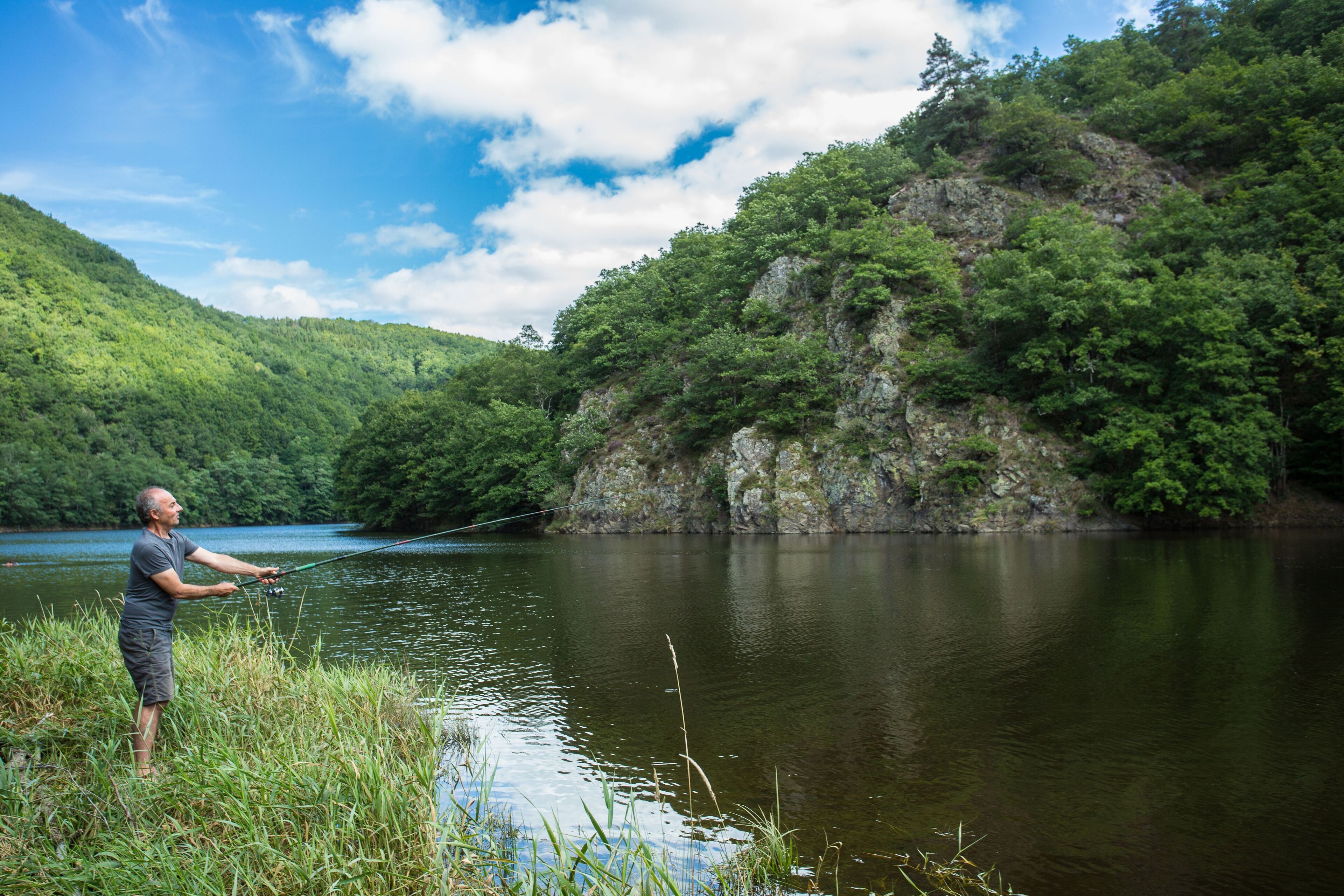 A man fishing by a calm lake surrounded by lush greenery and hills.