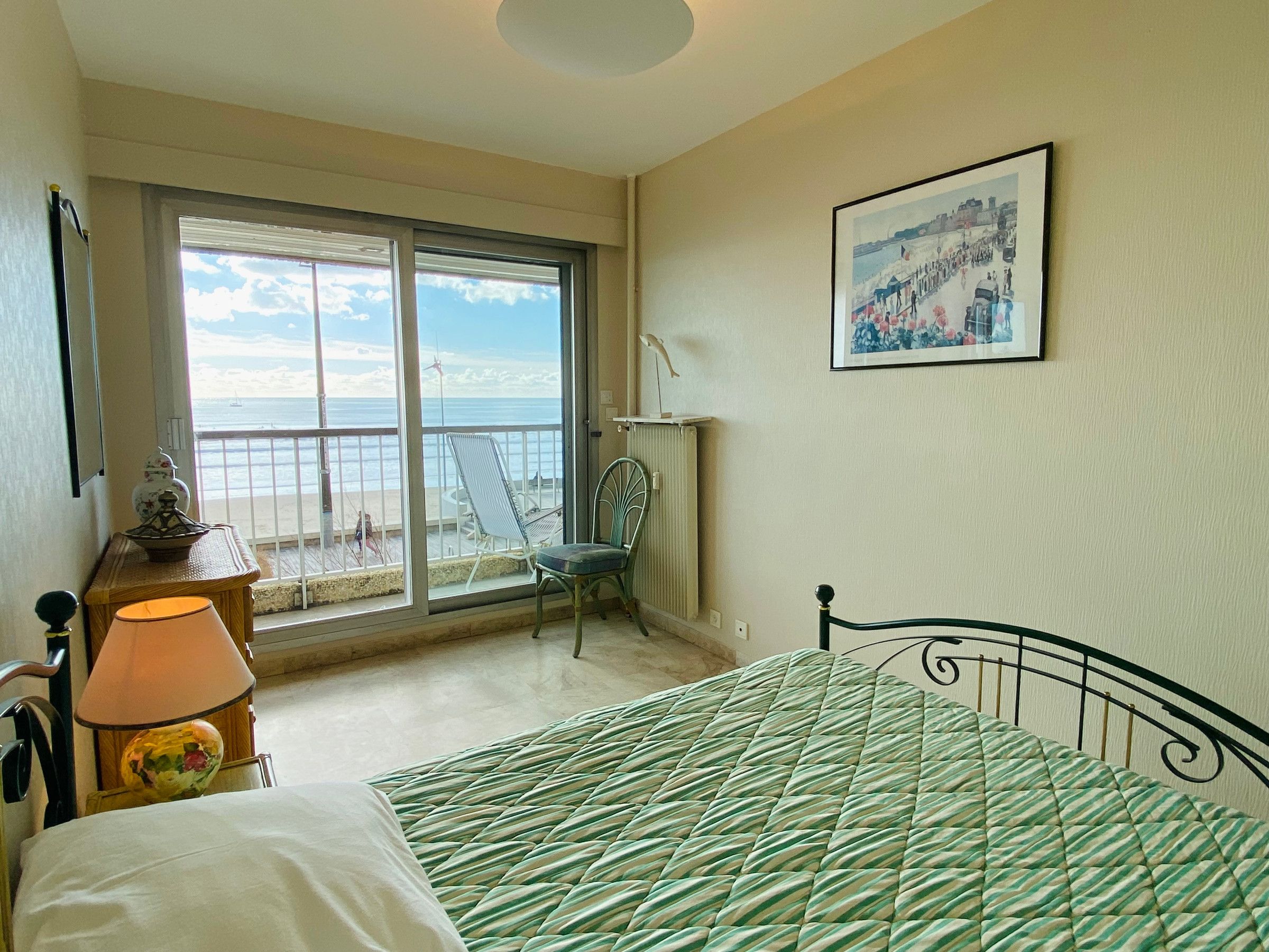 Une chambre à coucher avec un lit, une chaise, une lampe, un tableau au mur et une vue sur la plage par la fenêtre.