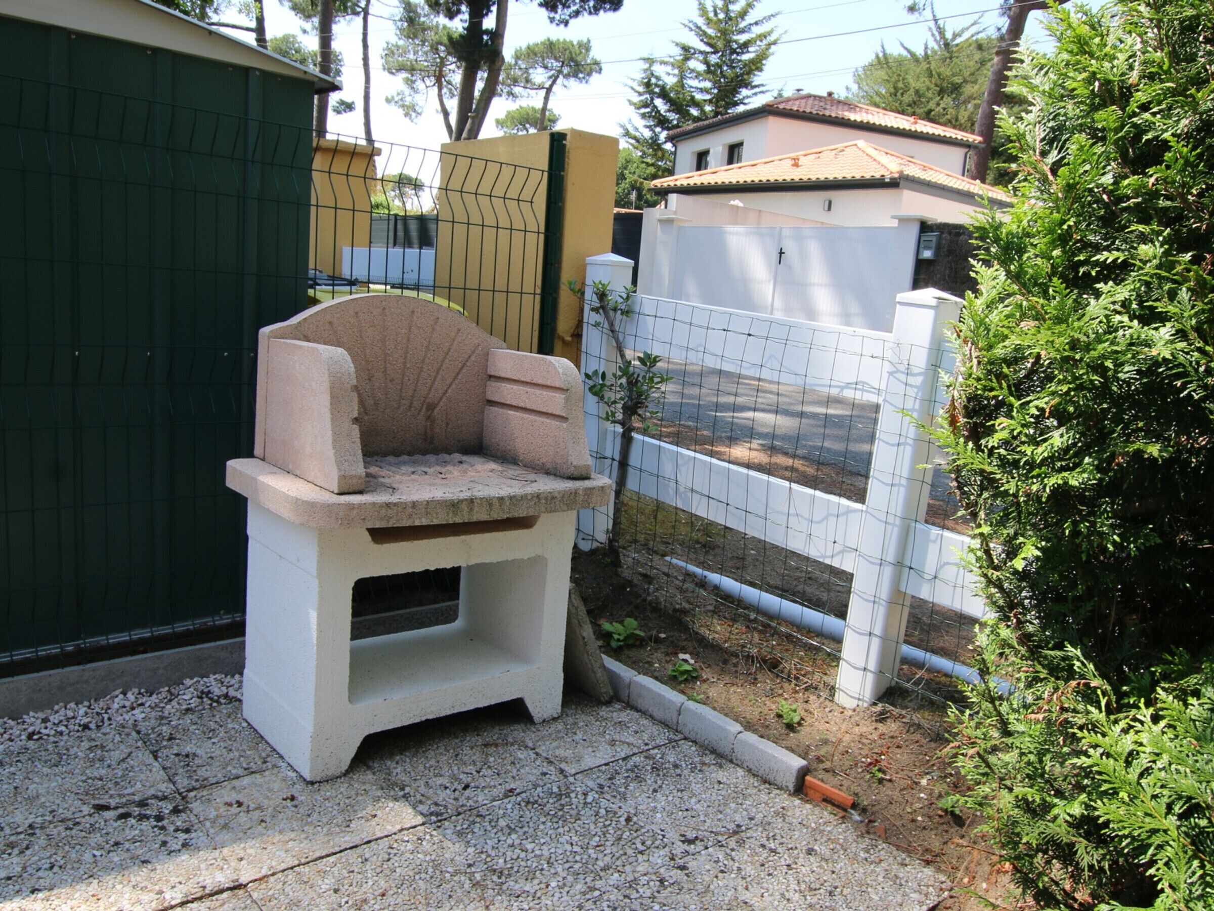 A concrete outdoor seating area with a stone bench, surrounded by a white fence and greenery.
