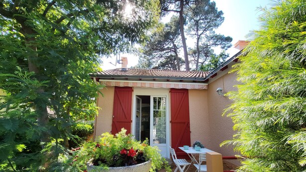 Eine kleine Terrasse mit Tisch und Stühlen, umgeben von viel Grün und einem Gebäude mit roter Tür.