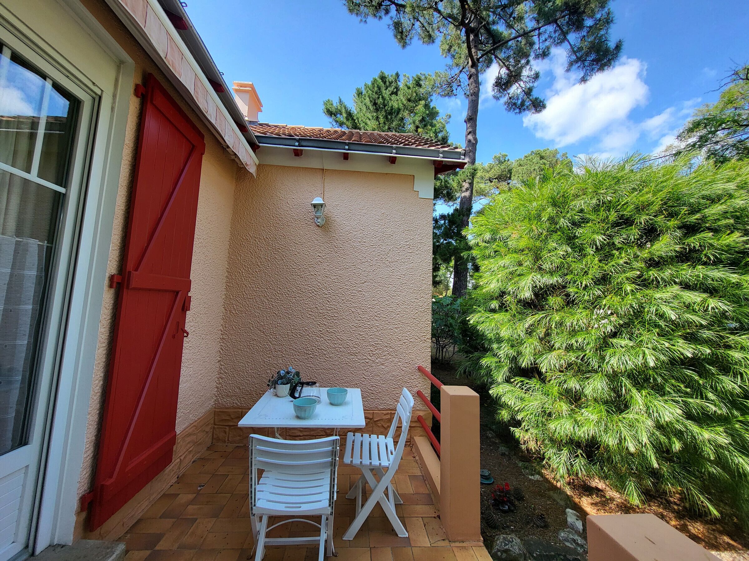 Eine kleine Terrasse mit Tisch und Stühlen, eine rote Tür und eine weiße Wand.