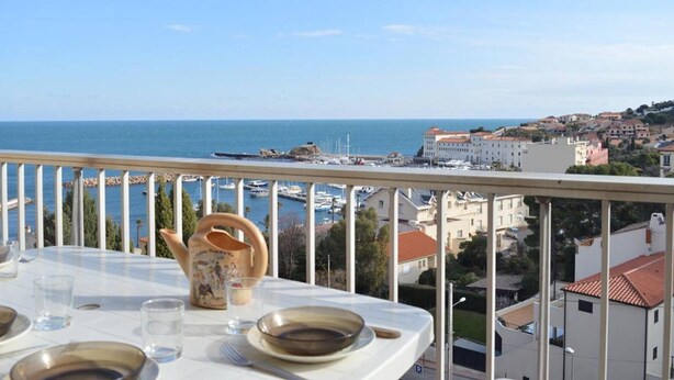 A balcony with a table set for dining, overlooking a marina and coastal town. - 2-bedroom apartment, sleeps 6, air conditioning and parking in Banyuls (Banyuls-sur-Mer)