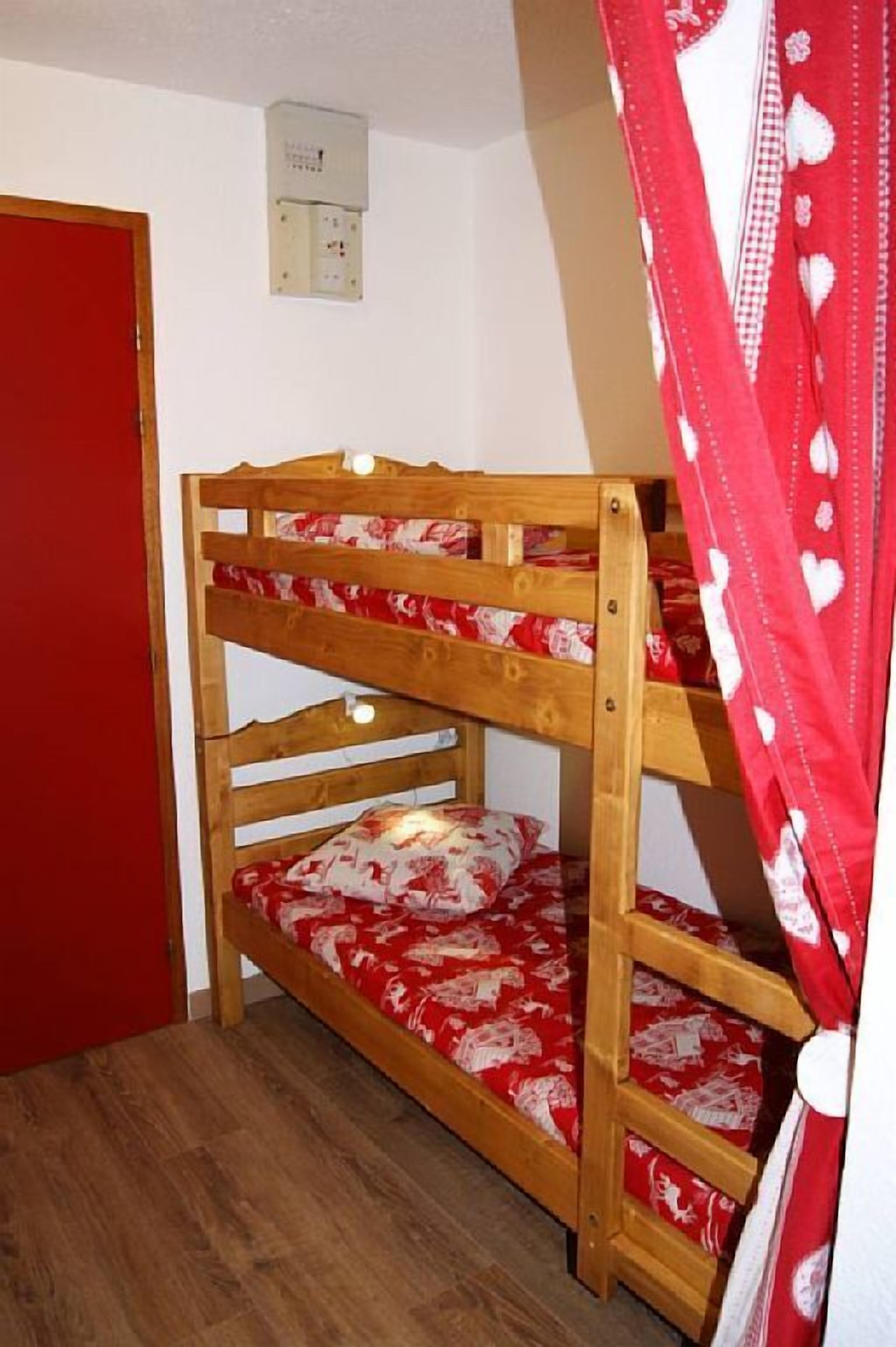 A bunk bed with red bedding, a wooden door, and a wall-mounted electrical box.