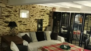 A cozy living room with a stone wall, a sofa set, a dining table with a checkered tablecloth, and a view through a glass door. - Gîte au Moulin (Loft) Pets welcome!  (Naucelle)