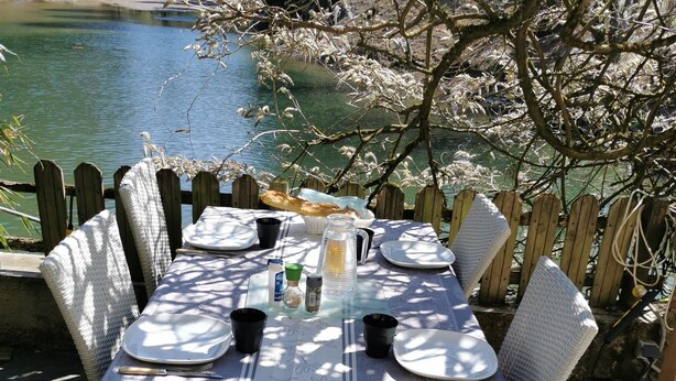 An outdoor dining table set with plates, cutlery, and a pitcher, surrounded by wicker chairs, with a view of a lake and trees in the background. - Gîte au Moulin (Loft) Pets welcome!  (Naucelle)