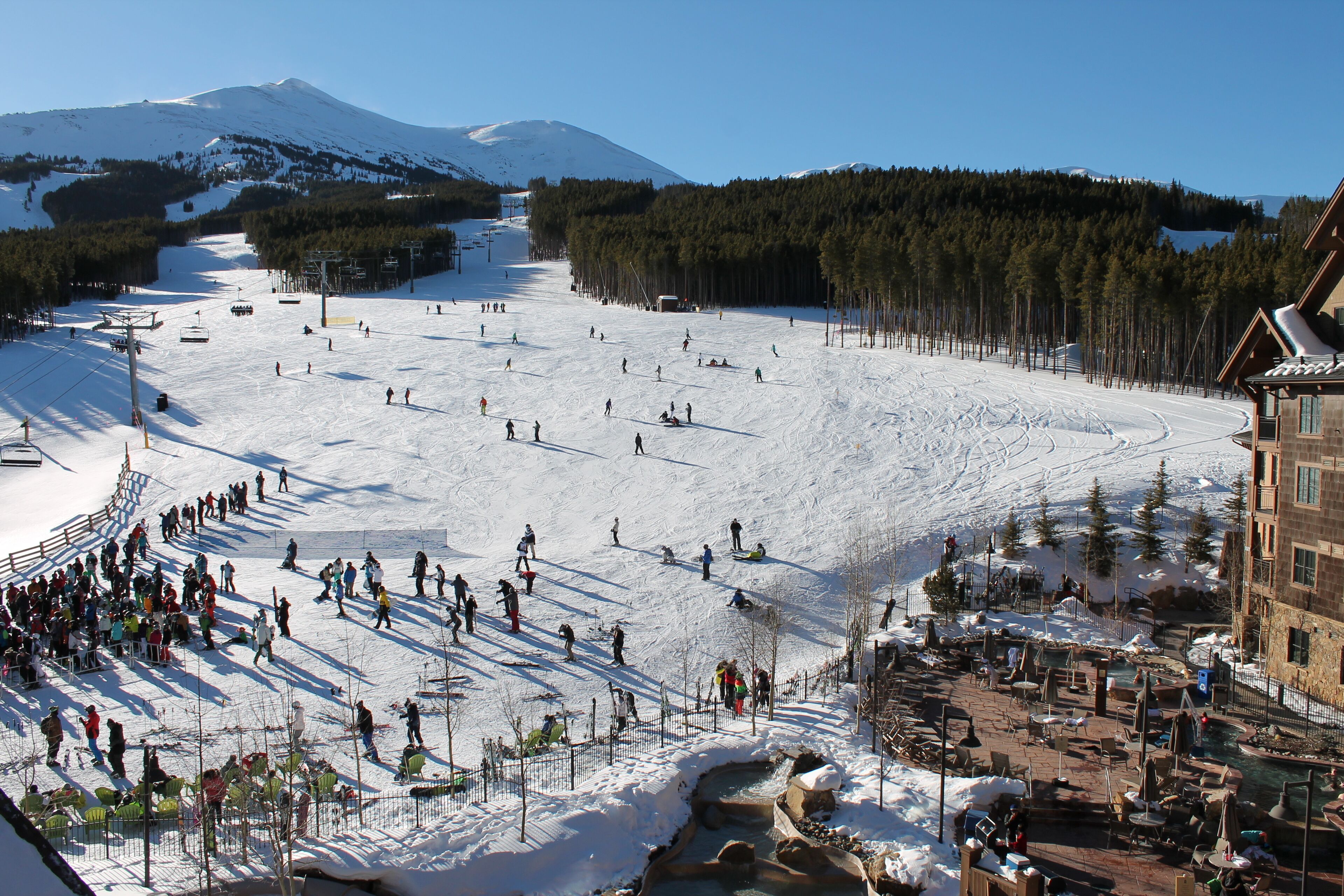 Ein Skigebiet mit einer weitläufigen Skipiste, einem Gebäude mit Seilbahn und einem Poolbereich.