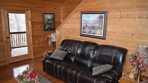 Interior de uma cabana de madeira com um sofá de couro, duas pinturas emolduradas, uma luminária e uma porta que dá para um deck.