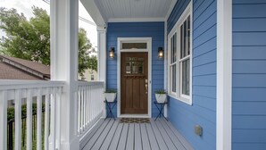 Uma casa azul com varanda branca, porta marrom e dois vasos de plantas de cada lado.