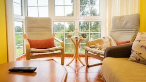 Une pièce avec deux chaises et un canapé, le tout placé face à une grande fenêtre donnant sur un paysage verdoyant. Une petite table avec une tasse et un livre se trouve au centre.