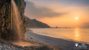 Un paysage côtier avec une cascade, une plage et des formations rocheuses lointaines, le tout baigné par les couleurs du coucher de soleil.