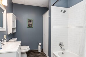 Een badkamer met een witte wastafel, toilet en douche. Er hangt een bordje in een lijst aan de muur en er ligt een rol wc-papier op de vloer.