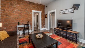 A living room with a brick wall, a ceiling fan, a TV on a wooden stand, a dining table with chairs, and a colorful rug. - Amazing historic home from 1860!  (St Charles)