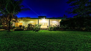 A modern house with a well-lit entrance, surrounded by a lush green lawn and palm trees. - BIG! 3BR Home Fully Equipped, Pool, Gardens & WiFi (El Cedral)