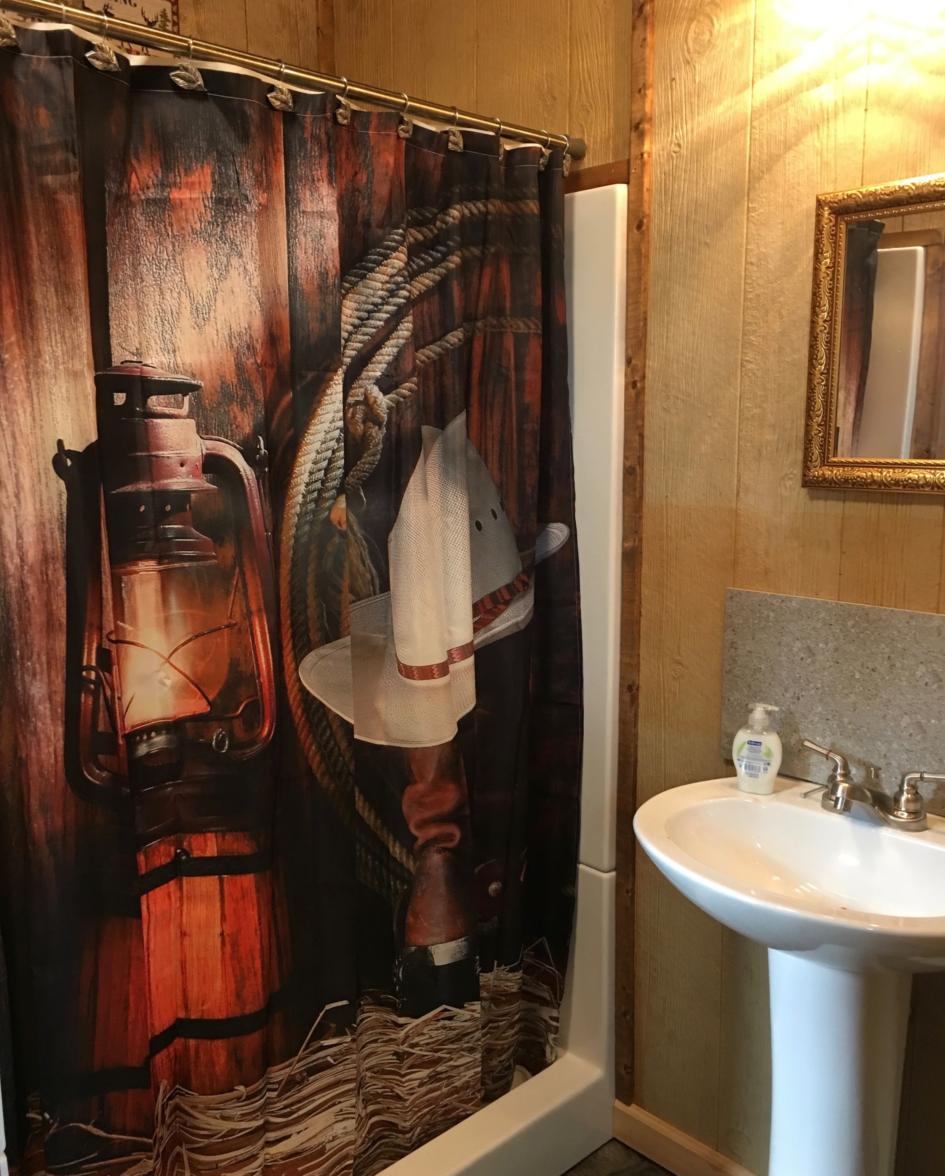Un baño con temática rústica de madera, que incluye una cortina de ducha con un farol y un balde antiguos, un lavabo blanco con espejo y una pared de madera.