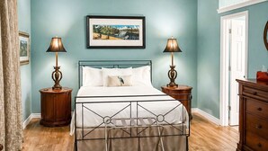 A bedroom with a bed, a ceiling fan, two bedside tables with lamps, a framed picture on the wall, and a wooden floor. - Centrally Located- 2 mi. North of downtown OS with screened porch, full kitchen (Ocean Springs)