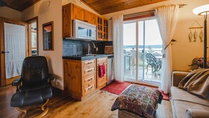 Ein gemütliches Zimmer mit Holzbalkendecke, einer Küchenzeile inklusive Mikrowelle, einem schwarzen Ledersessel, einem Sofa und Blick durch eine Glastür.
