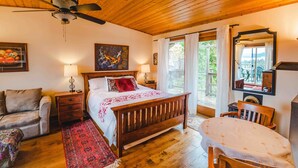 Ein Schlafzimmer mit Holzboden, einem Bett mit rotem und weißem Überwurf, einem Nachttisch aus Holz, einem Teppich mit rotem und weißem Muster, einem Holzstuhl, einem Tisch mit weißem Tuch sowie einem Deckenventilator und einem Fenster mit Vorhängen.