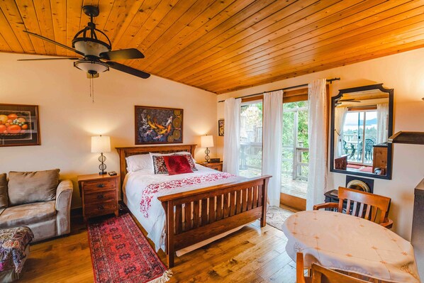 A bedroom with a wooden ceiling, a bed with a red and white bedspread, a wooden nightstand, a red and white patterned rug, a wooden chair, a table with a white tablecloth, a ceiling fan, and a window with curtains. - Morningside Garden Suite
FLASH SALE- CHECK OUT OUR WINTER GET AWAY SPECIAL (Salt Spring Island)