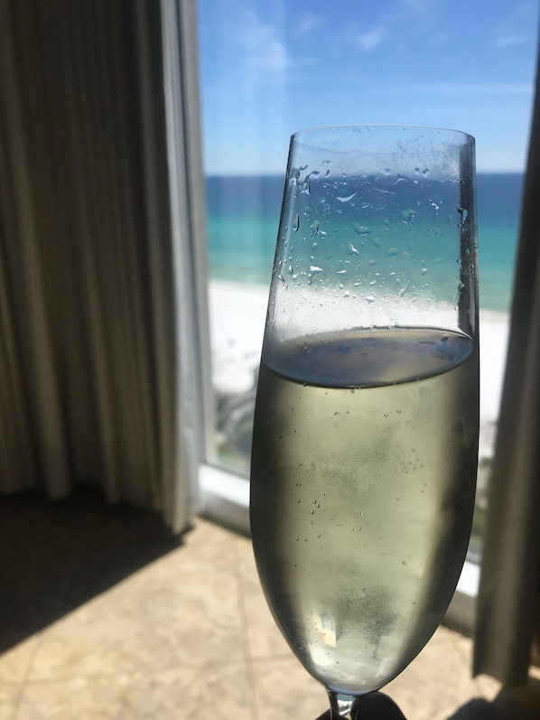 A glass of white wine on a windowsill with a view of the ocean and a curtain in the background.