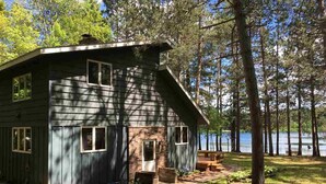 A rustic cabin with a large porch, a chimney, and a view of a lake surrounded by trees.