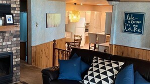 A cozy living room with a fireplace, a black leather sofa with blue cushions, wooden paneling on the ceiling, and a kitchen area in the background.