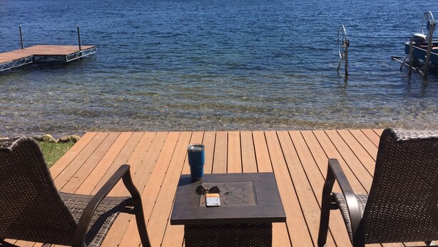 A wooden deck with two chairs and a table, overlooking a calm lake with a dock and trees in the background.