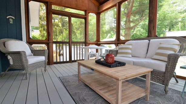 A covered outdoor seating area with wicker furniture, a wooden table, and a view of trees through large windows. - 4 Bedroom, Community Pool!  517 Doral Drive, Bethany West (Bethany Beach)
