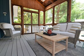 Un espace extérieur aménagé, abrité et doté de mobilier en osier, d’une table en bois et offrant une vue sur les arbres à travers de grandes fenêtres.