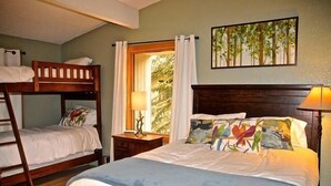 A bedroom with a bunk bed, a single bed, a nightstand with a lamp, a window with curtains, and a painting on the wall.