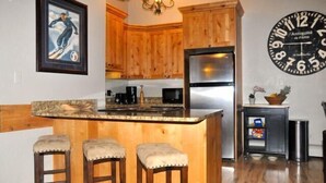 A kitchen with wooden cabinets, a stainless steel refrigerator, a bar area with stools, and a wall clock.