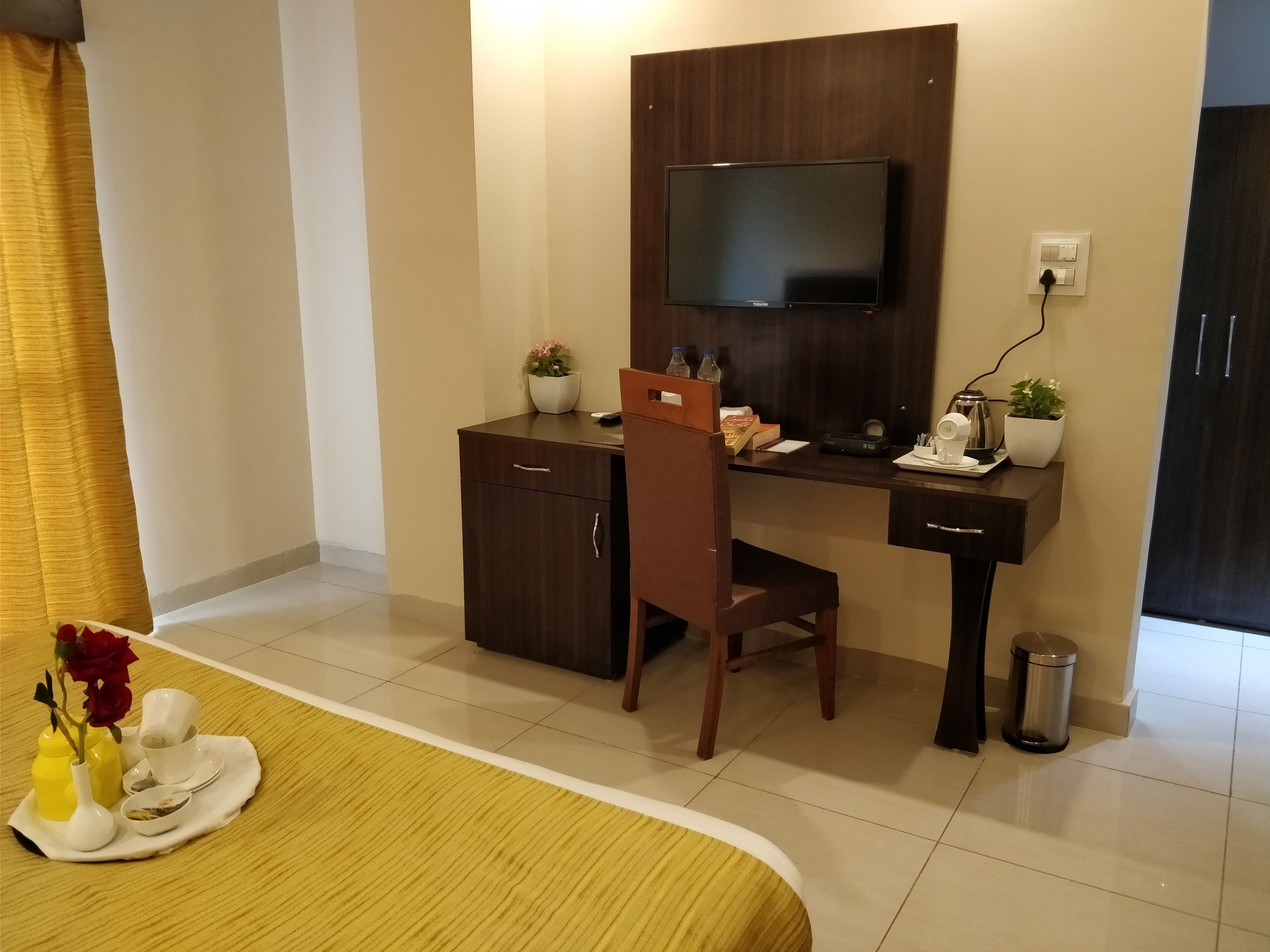 A hotel room with a bed, a desk with a flat-screen TV, a chair, a small trash bin, and a potted plant.