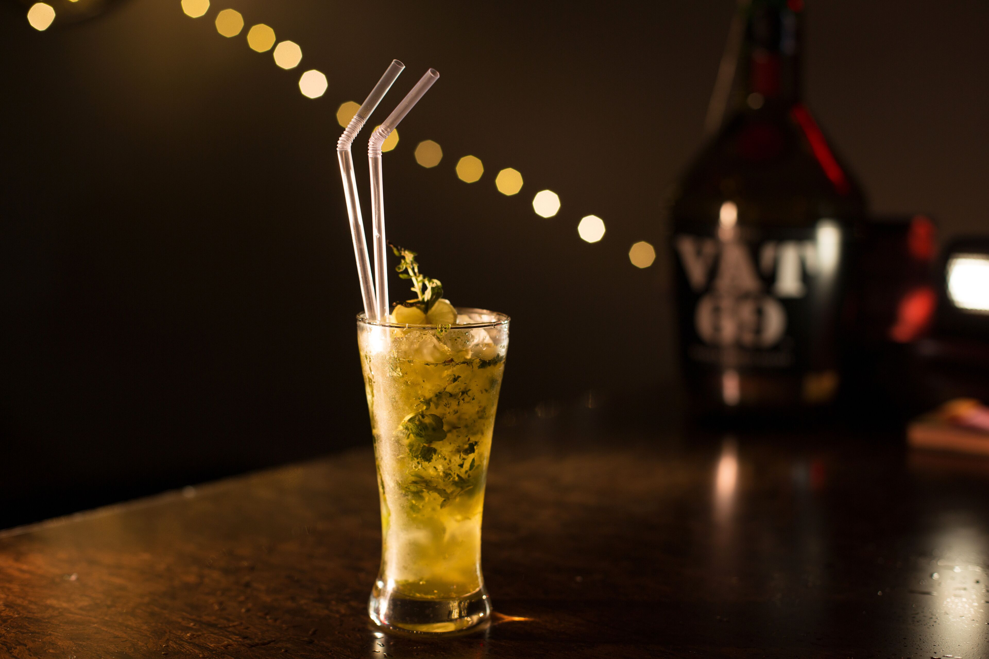 A glass of cocktail with a straw, garnished with a lemon slice, on a wooden bar counter.