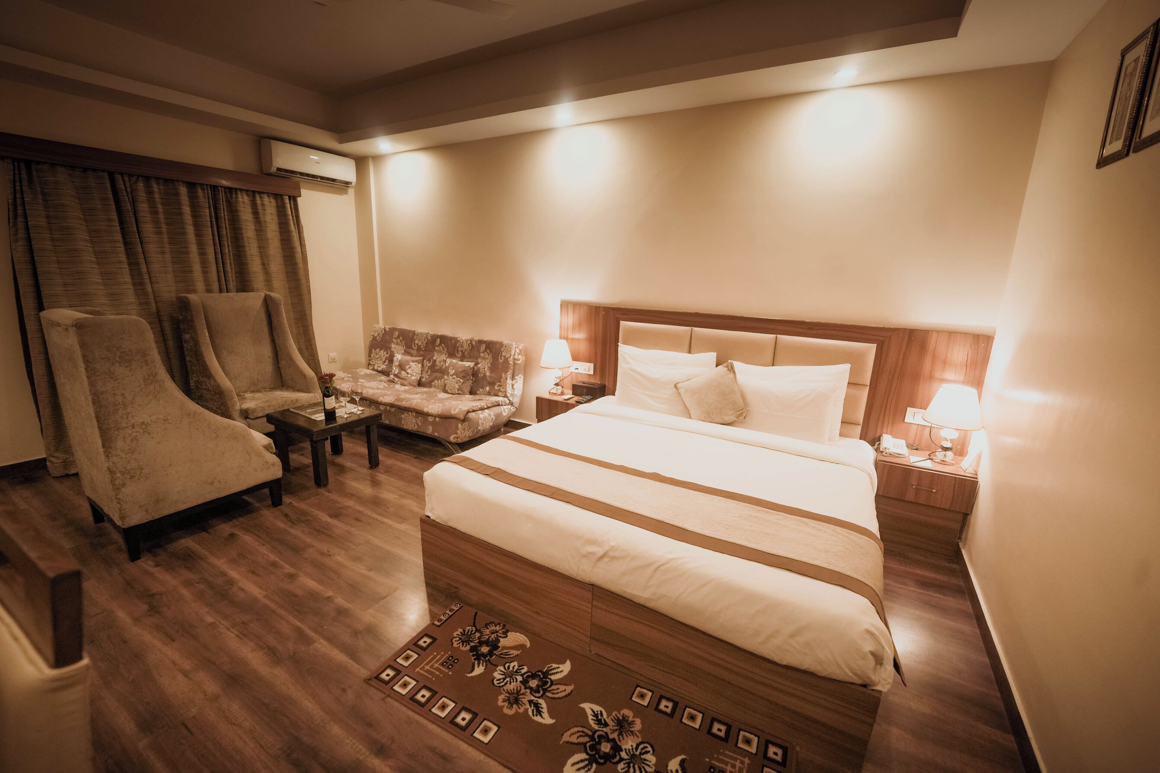 A hotel room with a large bed, two armchairs, a small sofa, a wooden floor, and a patterned rug.