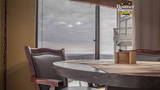 A round table with a woven placemat, two upholstered chairs, and a wooden side table with a candle. A large window offers a view of the outdoors. - Seven Springs Slope-Side Home, Ski right to the Lift #1  Boomerang Trail. (Champion)
