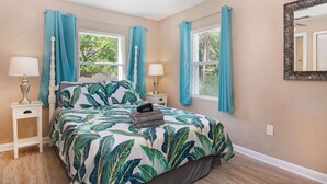 Une chambre à coucher comprenant un lit, deux tables de chevet, deux lampes, un ventilateur de plafond et deux fenêtres avec des rideaux.