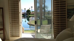A bedroom with a large bed, a sliding glass door leading to a balcony with a view of a lake and palm trees.