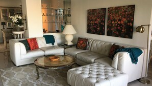 A modern living room with a white leather sectional sofa, a glass coffee table, and a patterned rug. There are two paintings on the wall and a floor lamp.