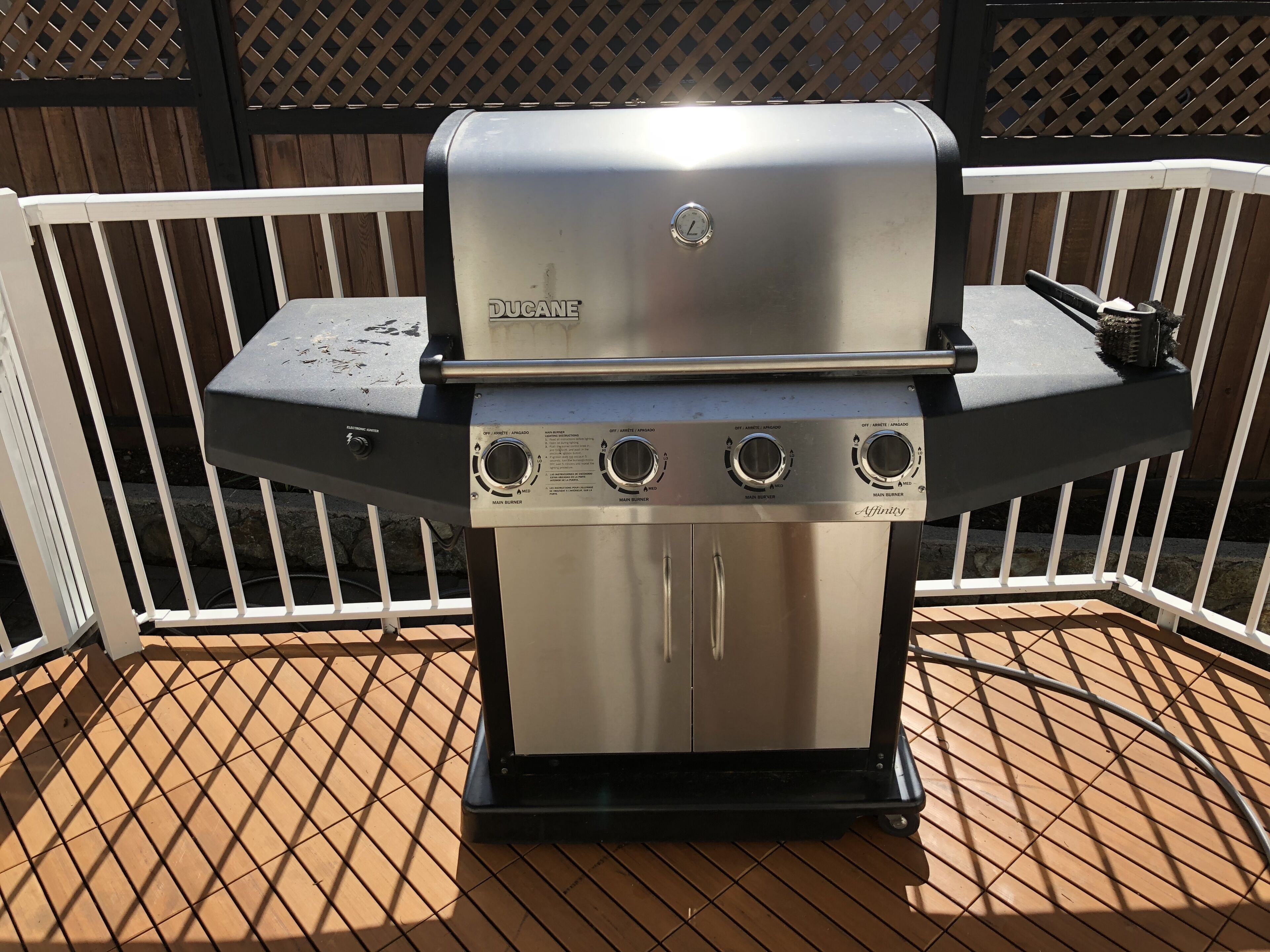 A stainless steel outdoor grill with four burners, a clock, and a control panel, set on a wooden deck with a white railing.