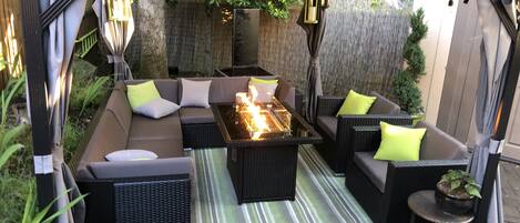 An outdoor patio with a sectional sofa, a glass-top table with a fire feature, wicker furniture, and a striped rug.