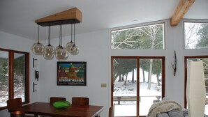 A dining area with a wooden table and chairs, a hanging light fixture, and a large window with a view of trees and a snowy landscape.